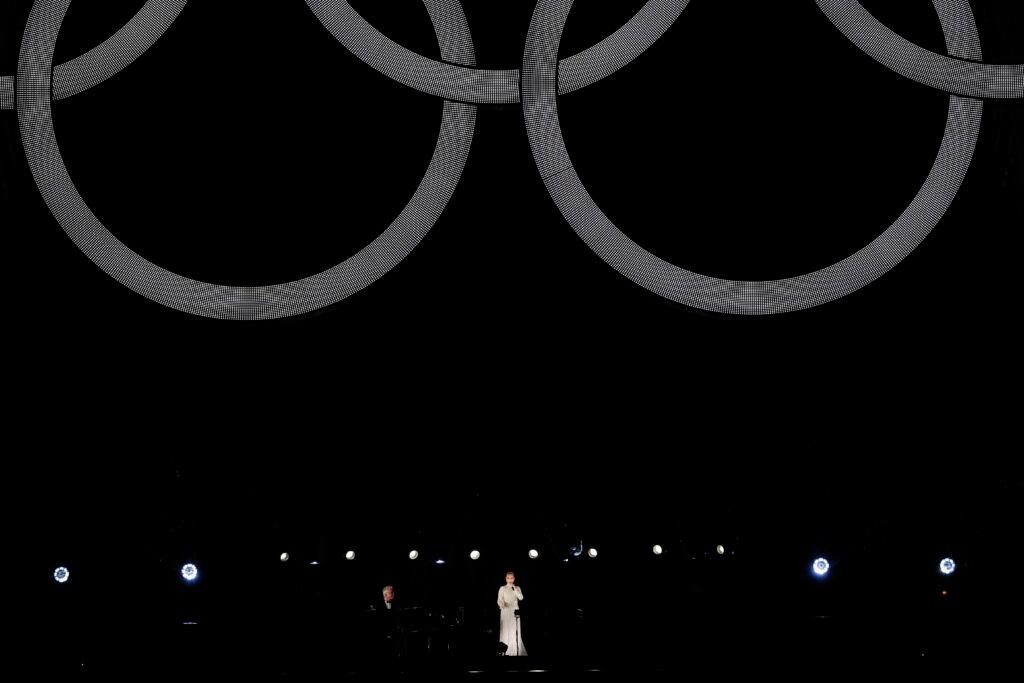 A wide shot of Celine Dion performing at the opening ceremony. The Olympic rings on the Eiffel Tower hang above her.