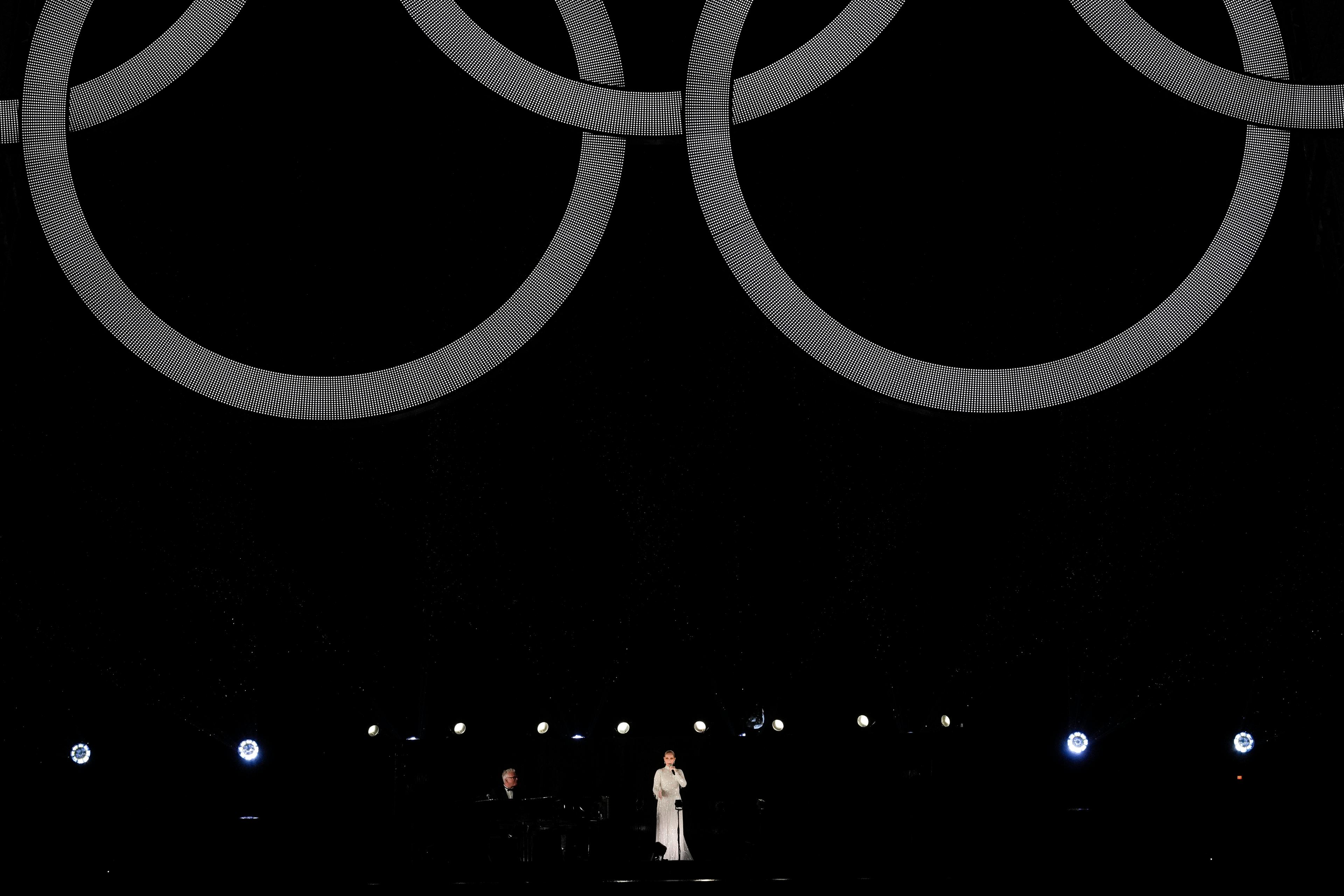 A wide shot of Celine Dion performing at the opening ceremony. The Olympic rings on the Eiffel Tower hang above her.