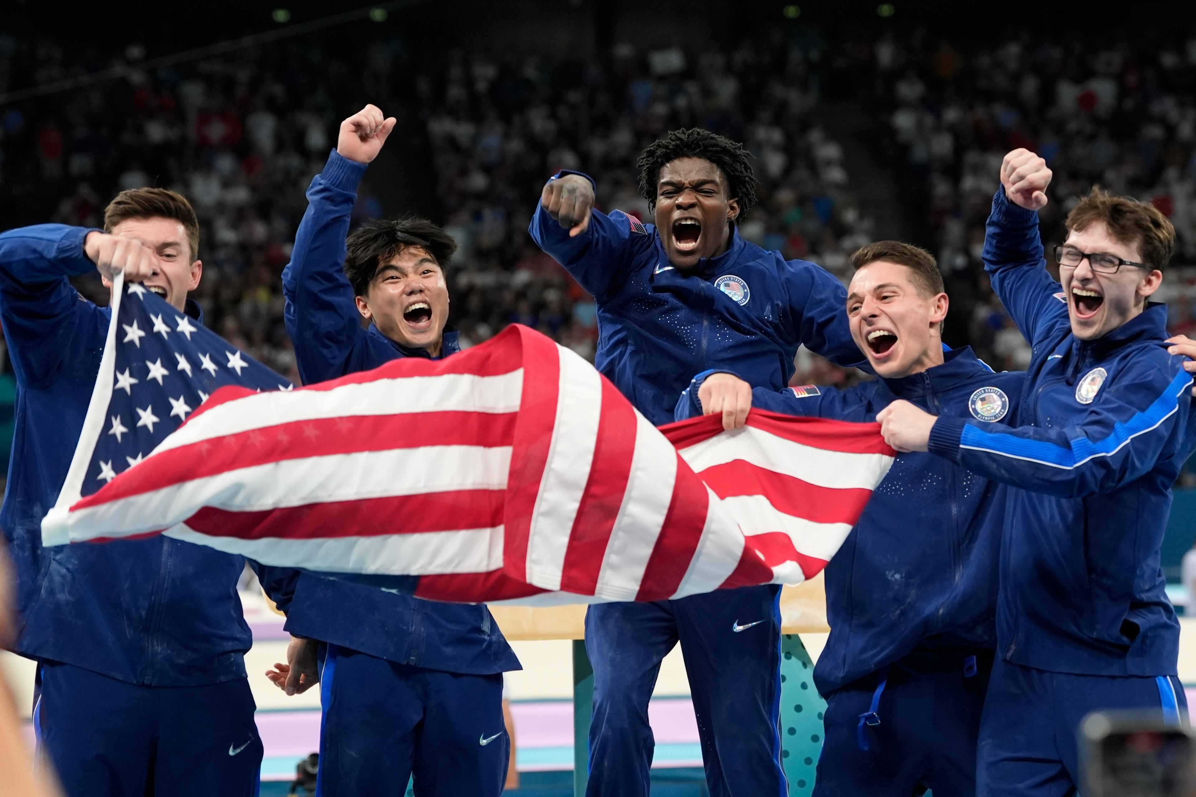 Five male USA gymnasts cheer while holding the American flag