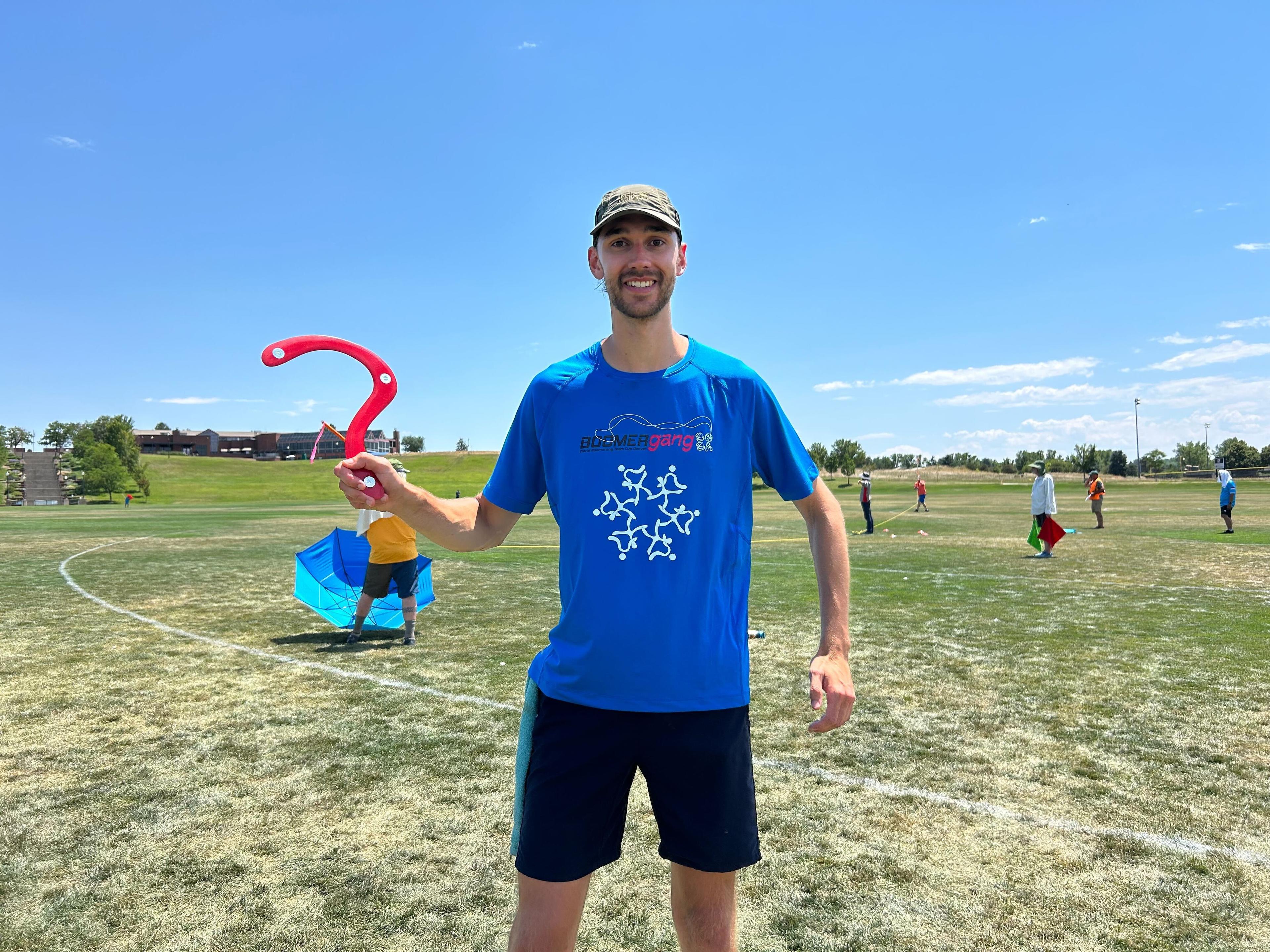 Man in blue shirt and grey cap poses with red boomerang he uses to make long distance shots.