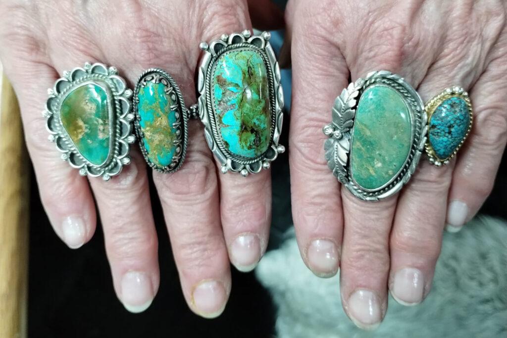 Five silver rings of different shapes and sizes feature large pieces of variegated blue and green turquoise. The rings are seen from the top and are won on a pair of hands. Three rings on the left hand and two on the right