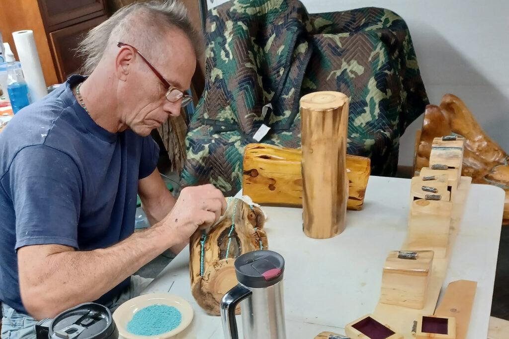 An older man with a light colored mohawk hairstyle and glasses wearing a dark blue t-shirt inlays small pieces of light blue turquoise into a polished piece of irregularly shaped wood. He's sitting at a work table with other wood pieces around him.