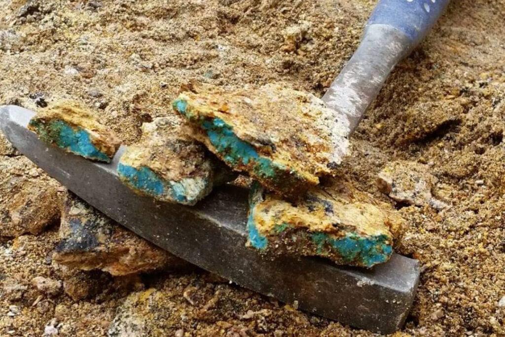 Four chunks of raw turquoise rest on the head of a hand tool used for digging that's laying on the ground.