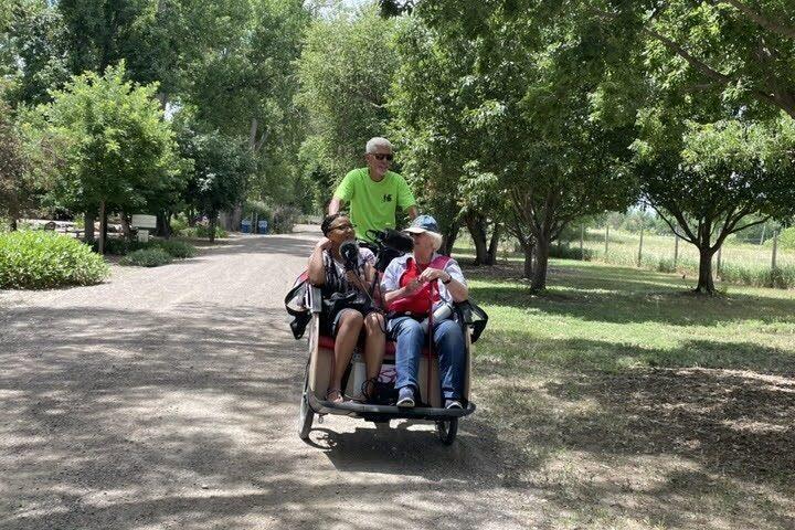 Reed Killam pedals a trishaw around Hudson Gardens with two passengers in front, Colorado Matters host Chandra Thomas Whitfield and Bonnie Douglas, who is celebrating her 85th birthday. There are microphones in front of Chandra and Bonnie to record their interview.