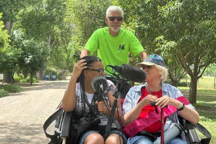 Reed Killam pedals a trishaw around Hudson Gardens with two passengers in front, Colorado Matters host Chandra Thomas Whitfield and Bonnie Douglas, who is celebrating her 85th birthday. There are microphones in front of Chandra and Bonnie to record their interview.