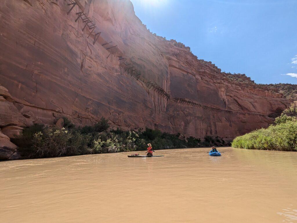 A packrafter and paddleboarder on the Dolores River observe the iconic Hanging Flume suspended over river.
