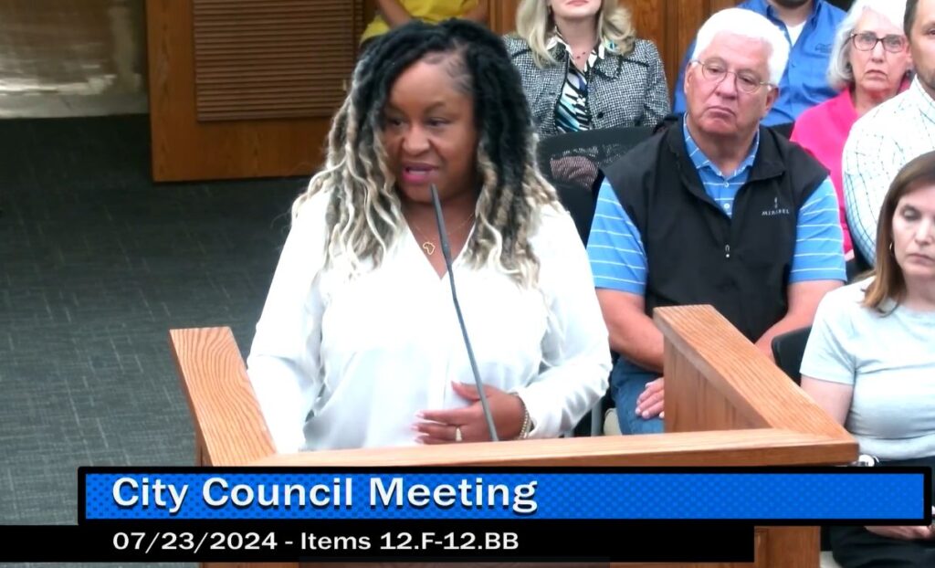 A woman of color with long long hair and wearing a white long sleeved shirt speaks into a thin microphone at a podium at a city council meeting. Behind her is a partial view of the other people attending the meeting.
