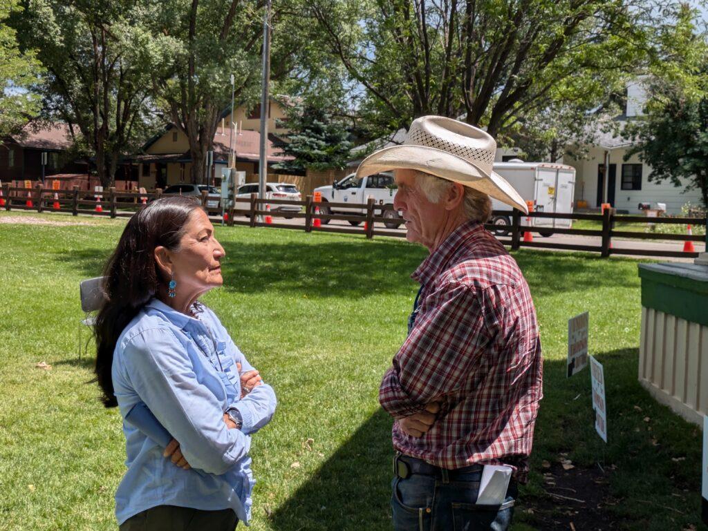 Deb Haaland standing to the left in a blue button down, speaks with rancher Bill Fales, standing to the right wearing a plaid button down shirt and a cowboy hat.