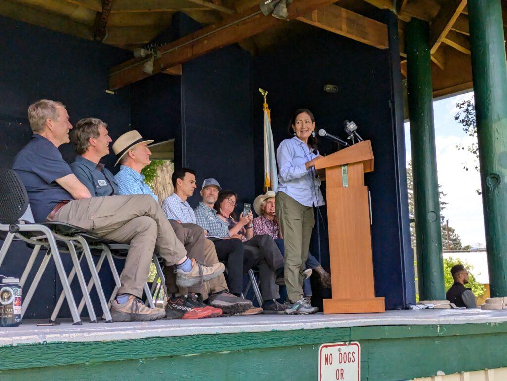 Interior Secretary Deb Haaland speaks at a podium wearing a blue button down shirt and green pants and sneakers while seven people sit next and behind her in chairs also on stage.