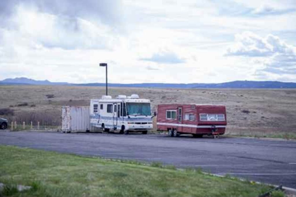 Two RVs sit off the side of a road near a light pole with a field and mountains in the background. One RV is a traditional-looing motor home. The other is smaller and red.