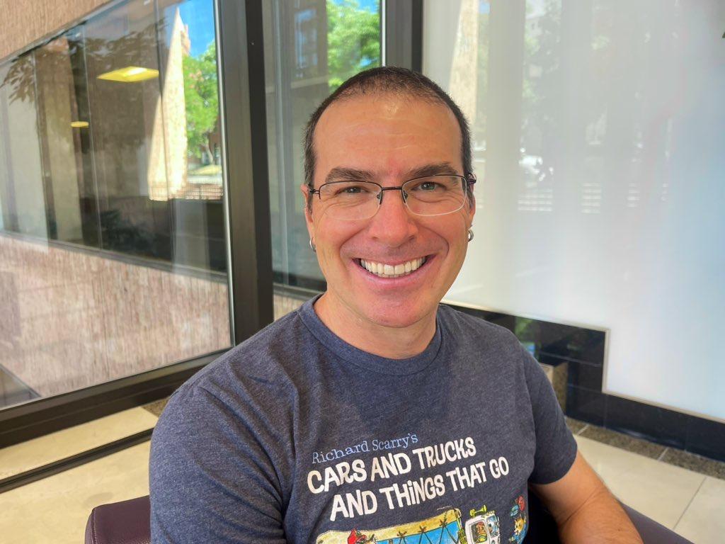 Paolo Bacigalupi sits in the CPR News lobby wearing a grey-blue t-shirt with glasses smiling at the camera.