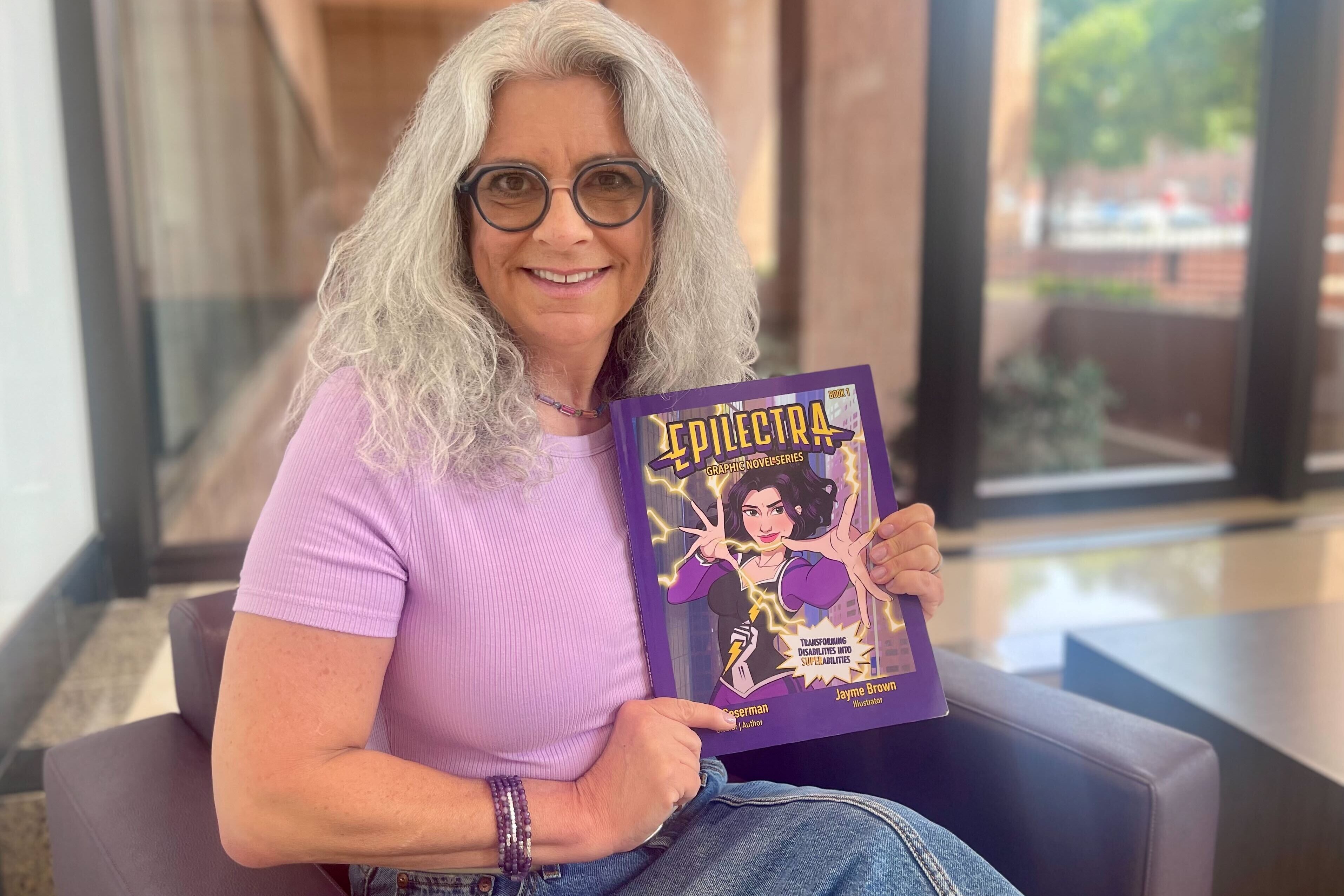A woman with white shoulder-length hair and a purple shirt holds up a comic book to her chest.