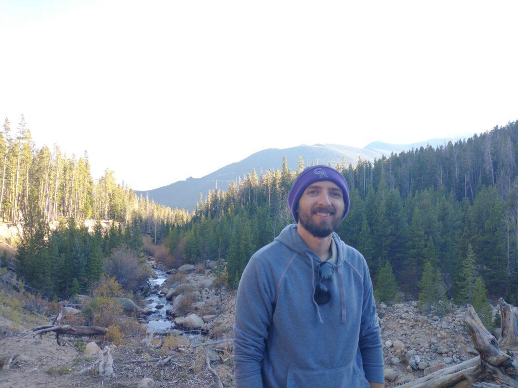 A man with a beard and a blue hoodie wearing a purple beanie with glasses attached to the front of his hoodie smiels at the camera with a small creek and trees and a mountain peak behind him.