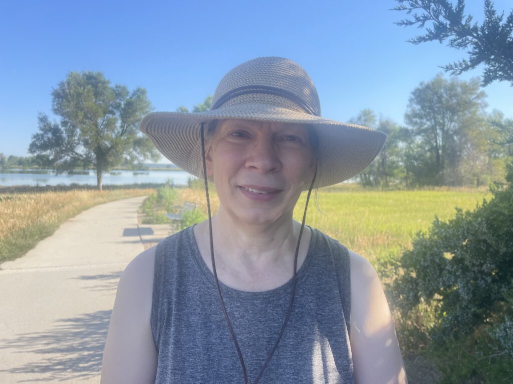 An older woman in a large straw hat with a walking trail and a lake in the background.
