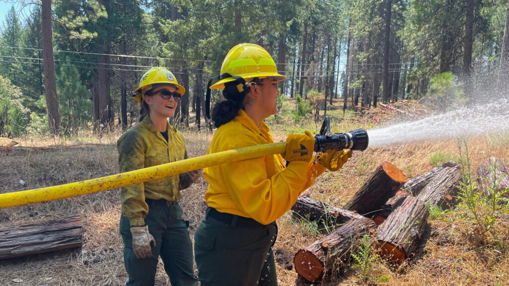 Two women wearing a yellow hard hat wearing long sleeve shirts and gloves holding a hose spraying water in the forest.
