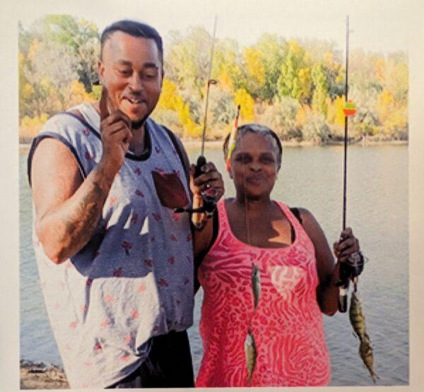 Kilyn Lewis stands with his mother fishing in front of a body of water with trees in the distance. His mother stands to his left holding a fishing pole.