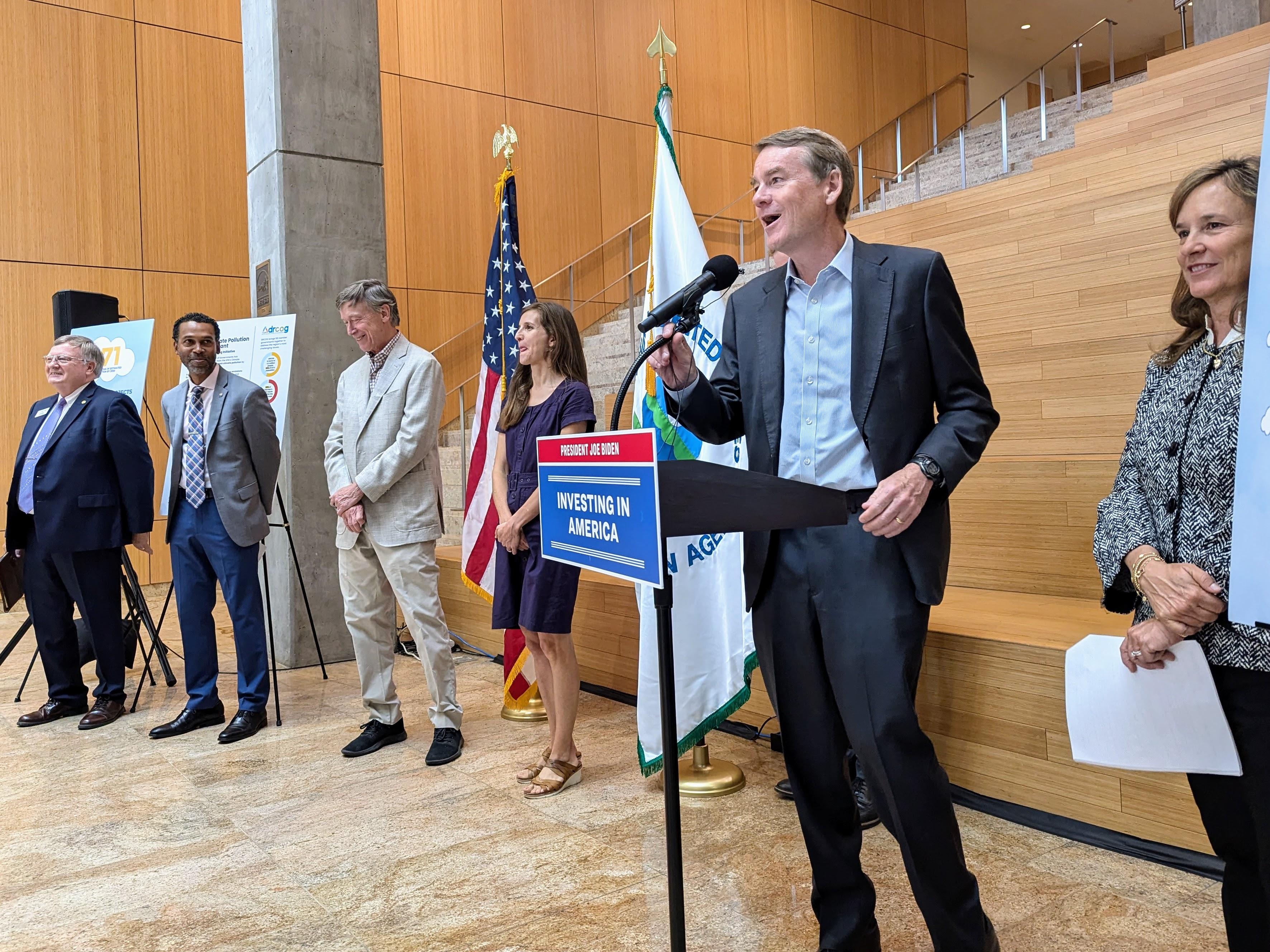 Six people stand in front of a series of stairs with two flags behind them on poles. One person, Michael Bennet, stands at a podium speaking into a microphone.