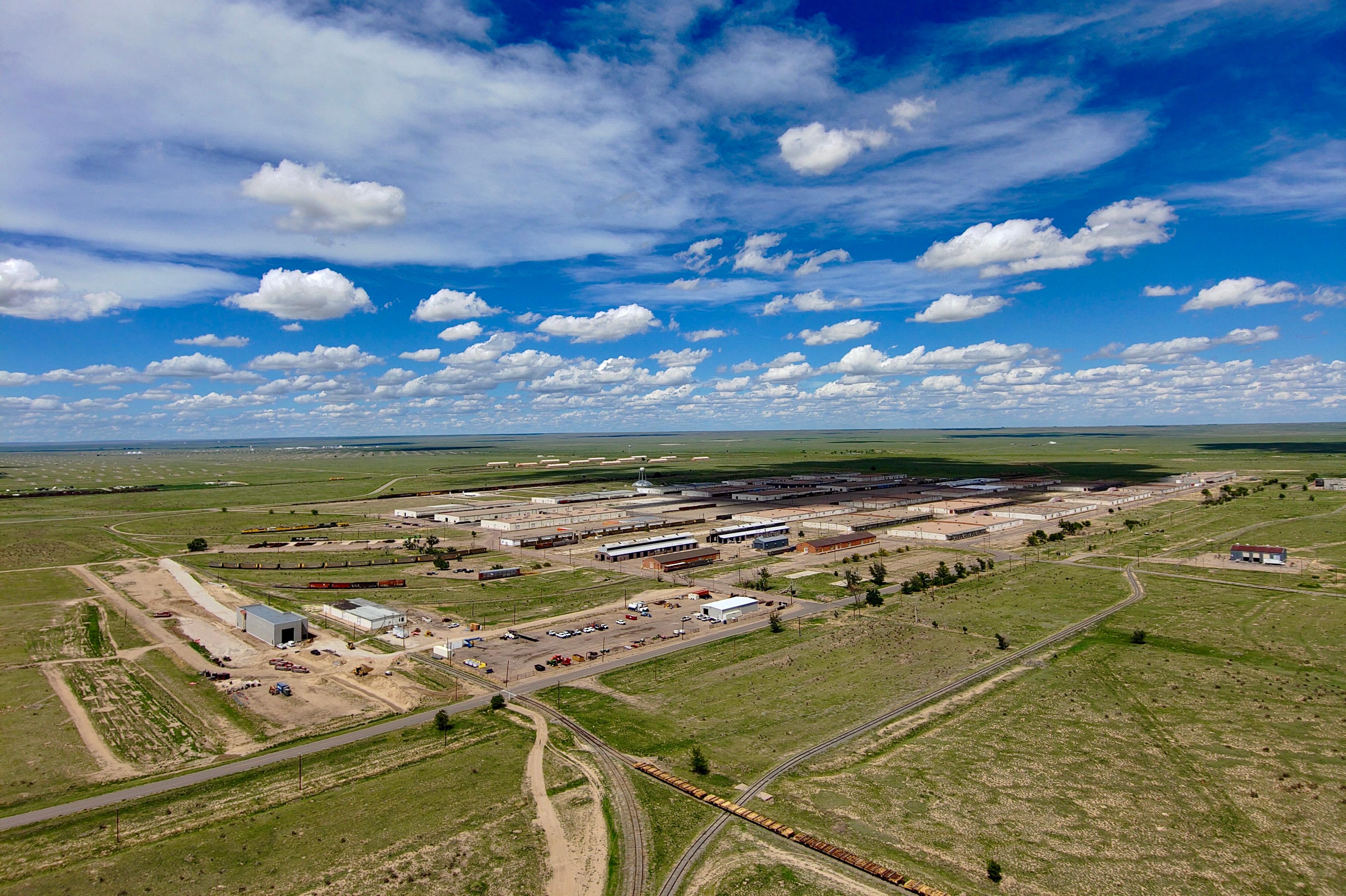An oblique aerial view of part of PuebloPlex showing a flat landscape with sparse green vegetation and several dozen long low industrial, warehouse and storage buildings lined up in rows along with a parking lot at the center of the image. More low buildings can be seen in the distance. Several roads lead into and through the area. The sky is blue with puffy white clouds.