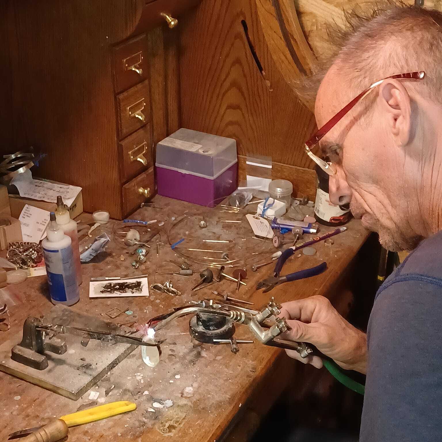 A man wearing glasses uses a torch at a workbench covered with other tools of a jewelers trade like pliers, tweezers, findings and more