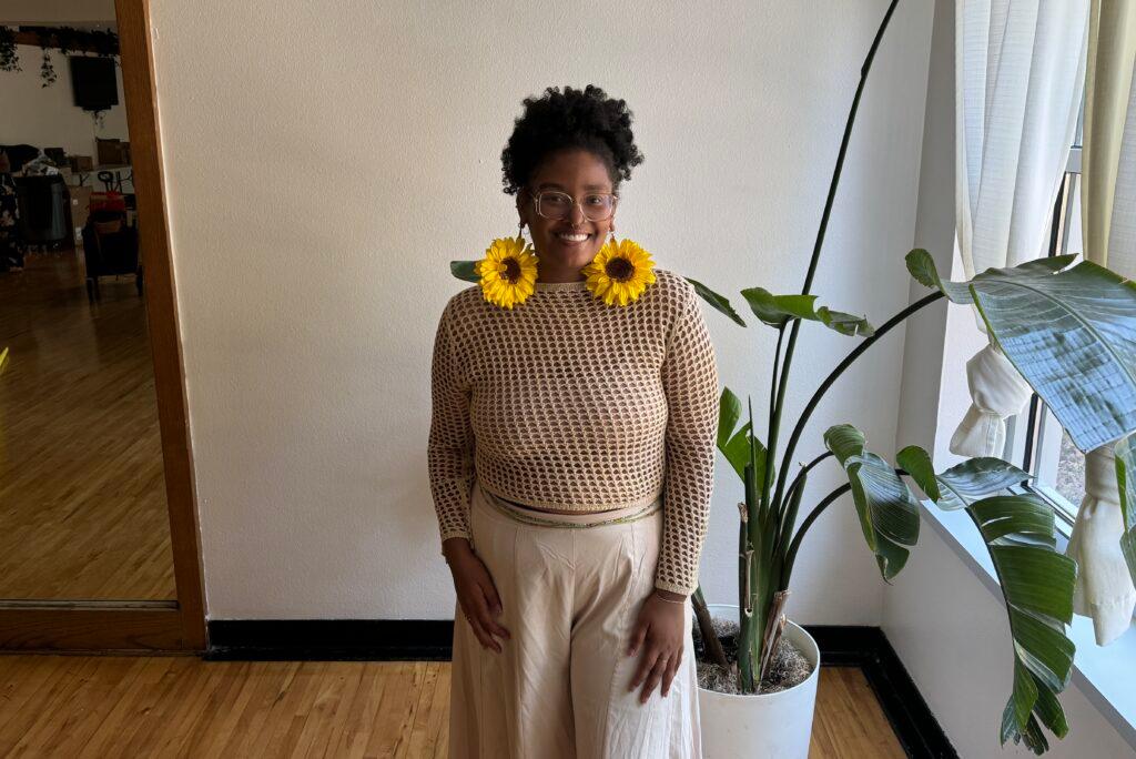 A woman poses with sunflower earrings in front of a potted plant.