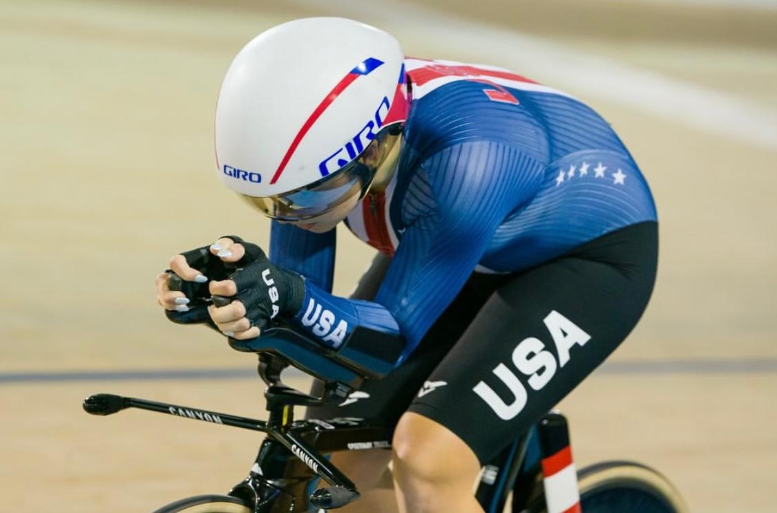 A cyclist in a blue kit wearing an aerodynamic white helmet with blut-tinted visor and Team USA shorts rides from left-to-right on a wooden velodrome track while.
