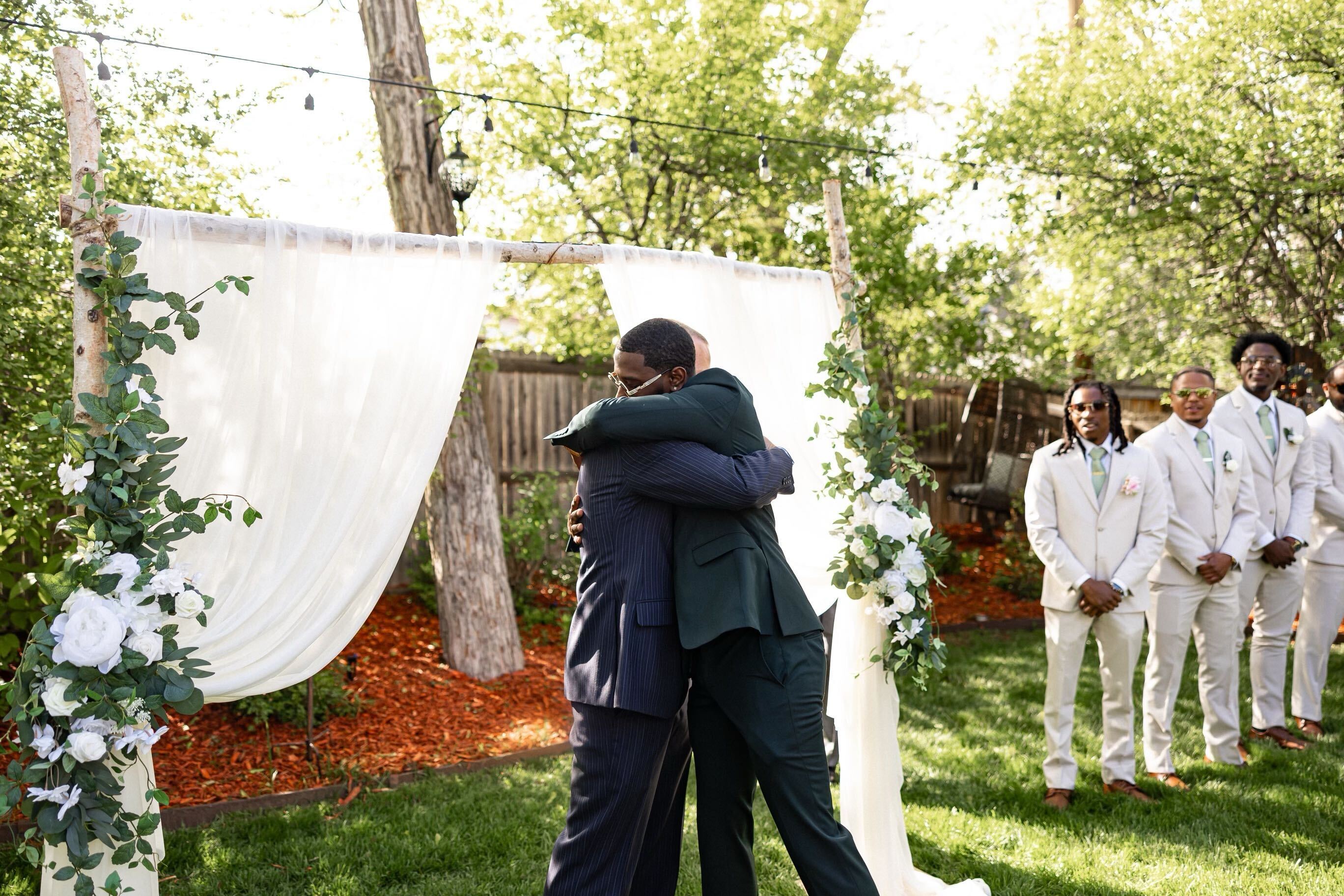 Two men embrace while a group of groomsmen stand nearby at a wedding.