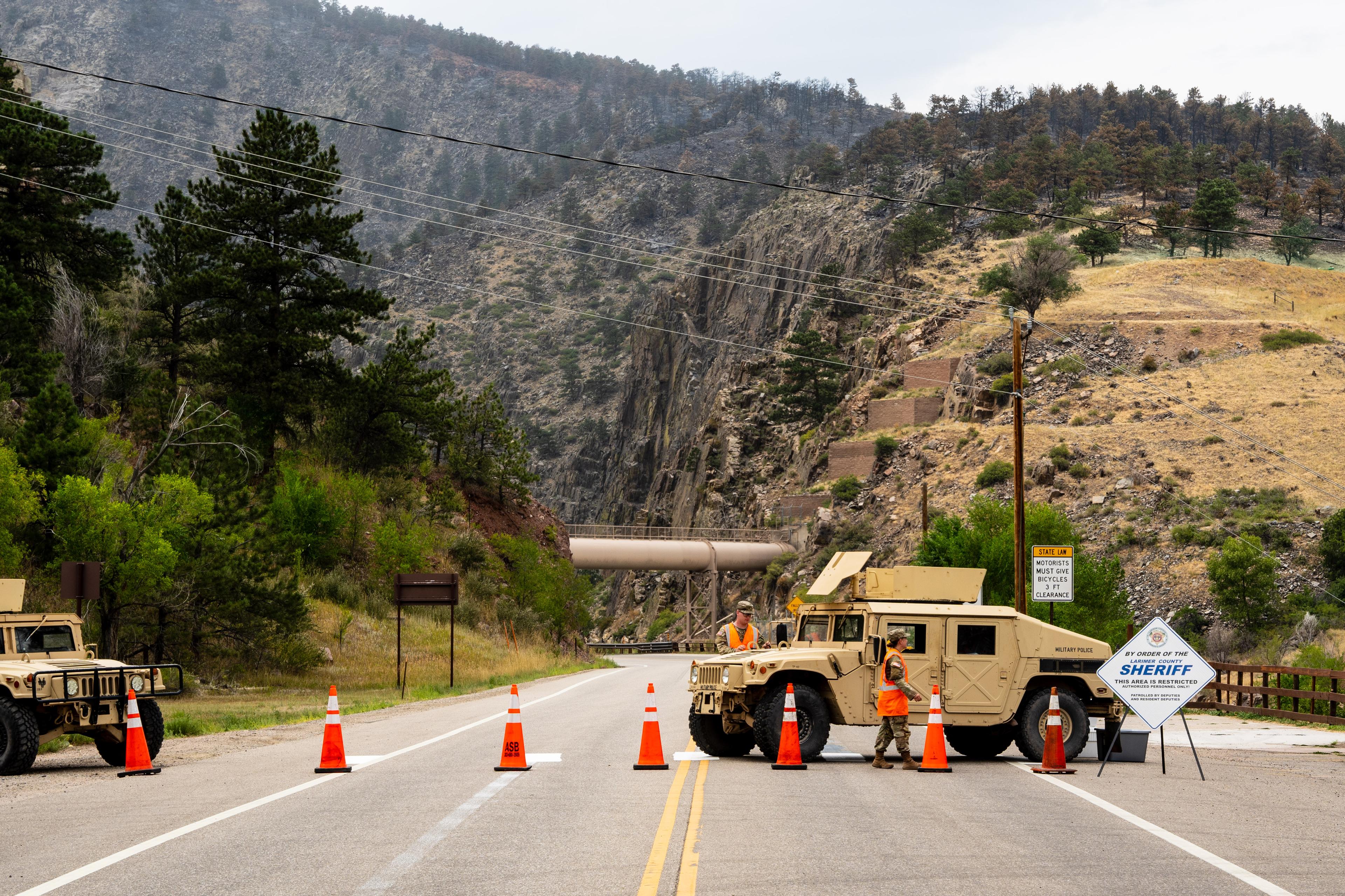 National guard troops block a road near a wildfire with a humvee and orange cones.