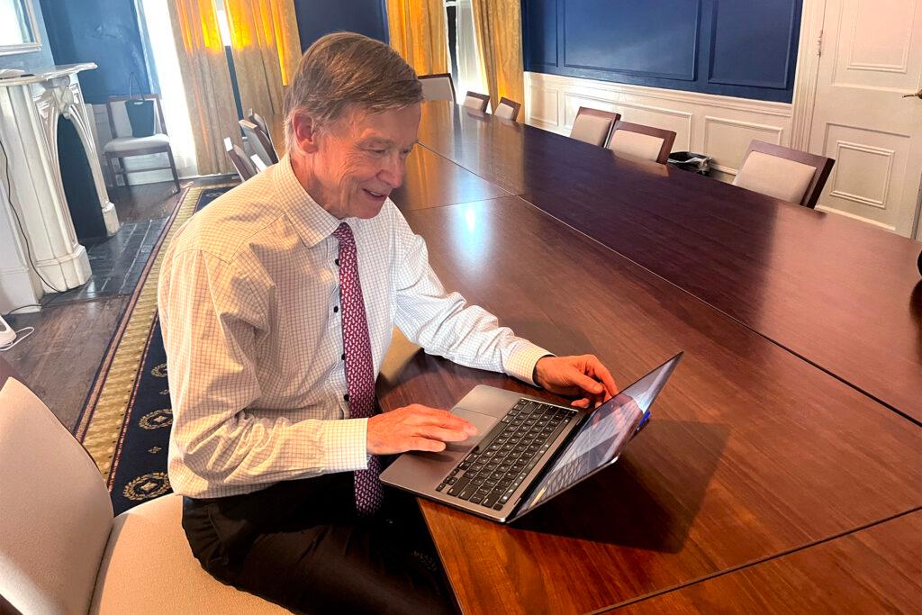 A man in a white checkered button up and a patterned red tie smiles at an open laptop, sitting in a fancy looking boardroom with a mahogany table and a fireplace.