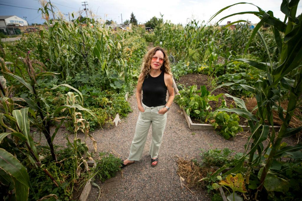 A woman in shades and a black tank top stands on a dirt path surrounded by overflowing garden plots. The scene around her is astoundingly green.