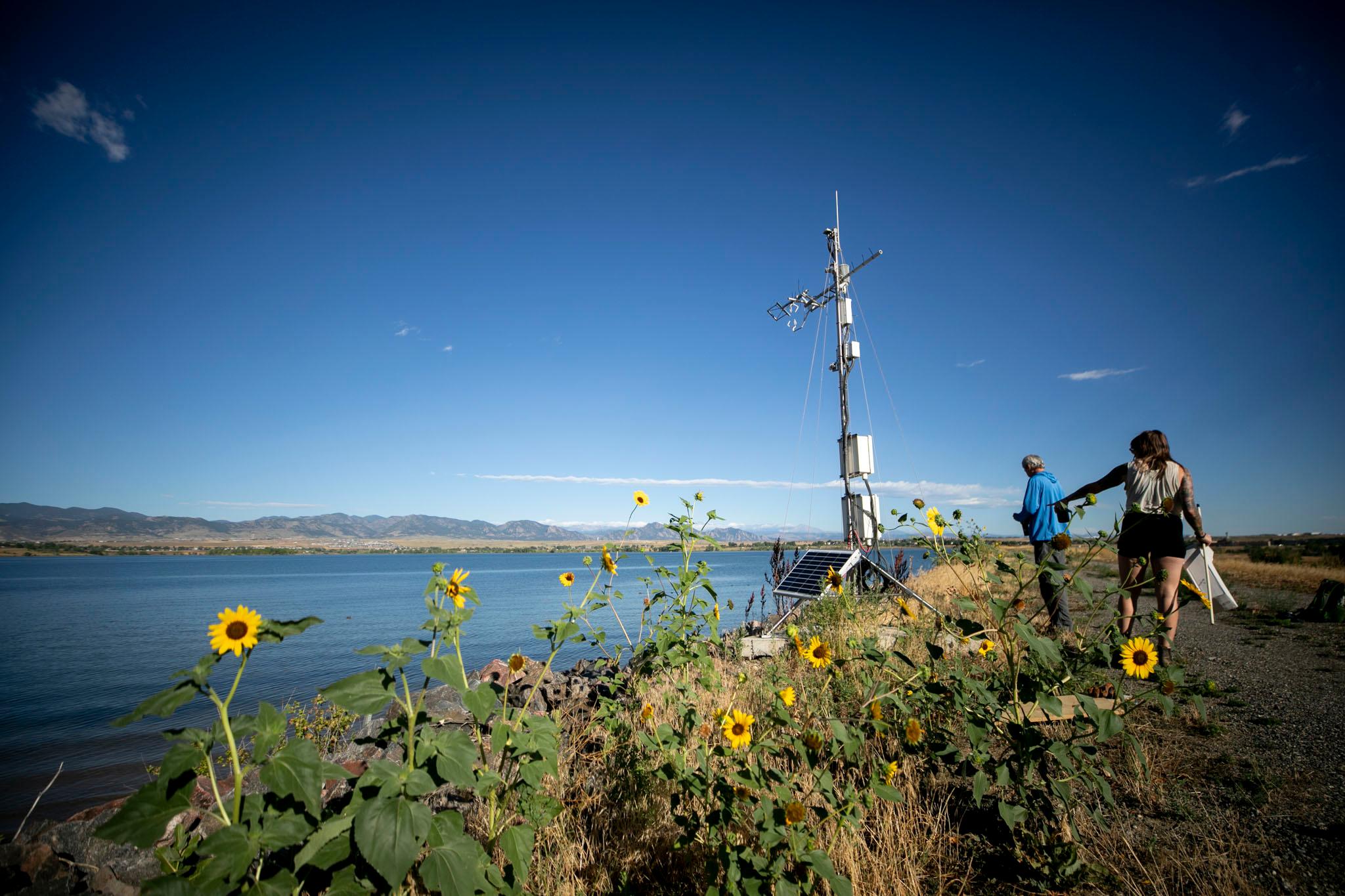 Two people stand on a ridge, lined with yellow flowers, above a blue lake and beneath blue skies; they're pointing at a tower of metal installed on the hill.