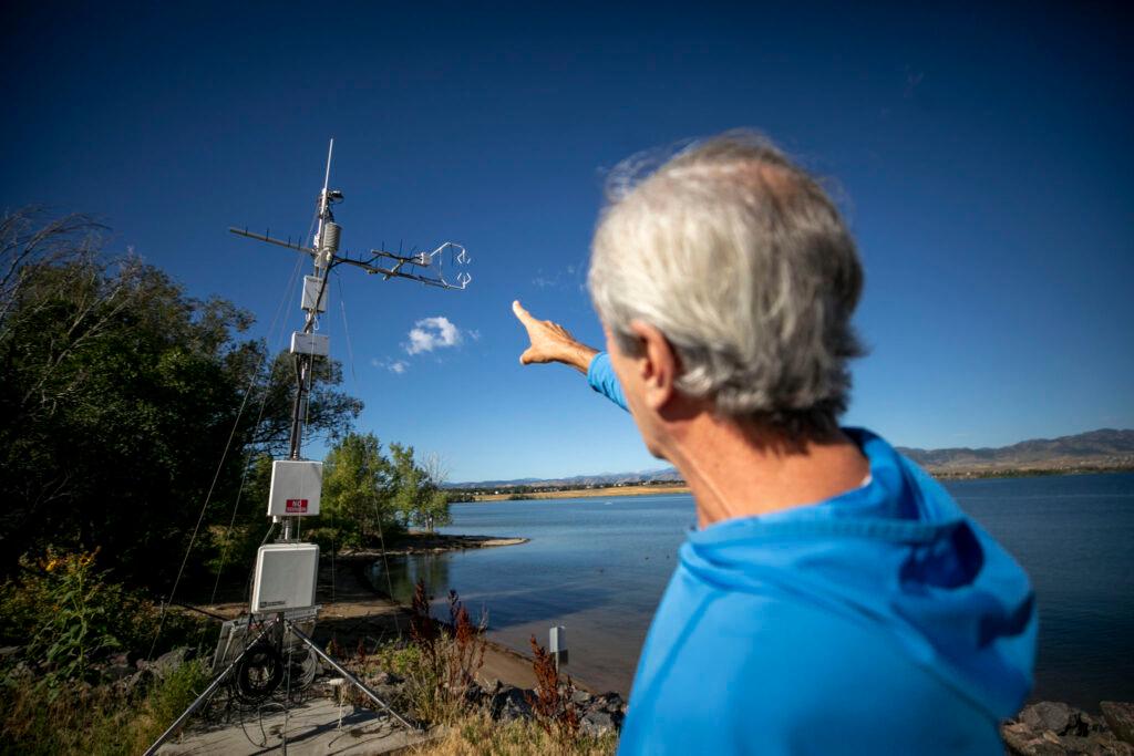 A man in a blue jacket points to a tower of metal instruments mounted on a green hill above a blue lake.