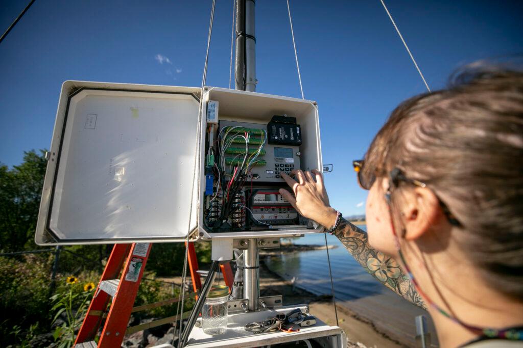 A view over a woman's shoulder, as her hands reach into a metal box filled with wires and electronics, mounted to a metal pole above a blue lake with a green shoreline.