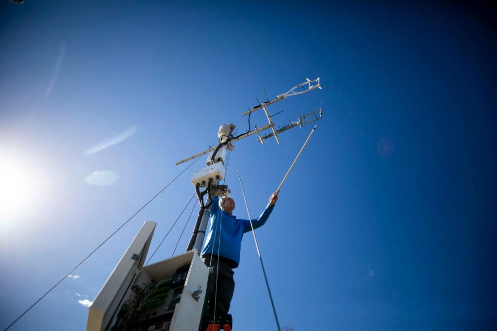 The camera looks up at a man standing beneath a blue sky, reaching up to a tower covered with electronic equipment with a stick.