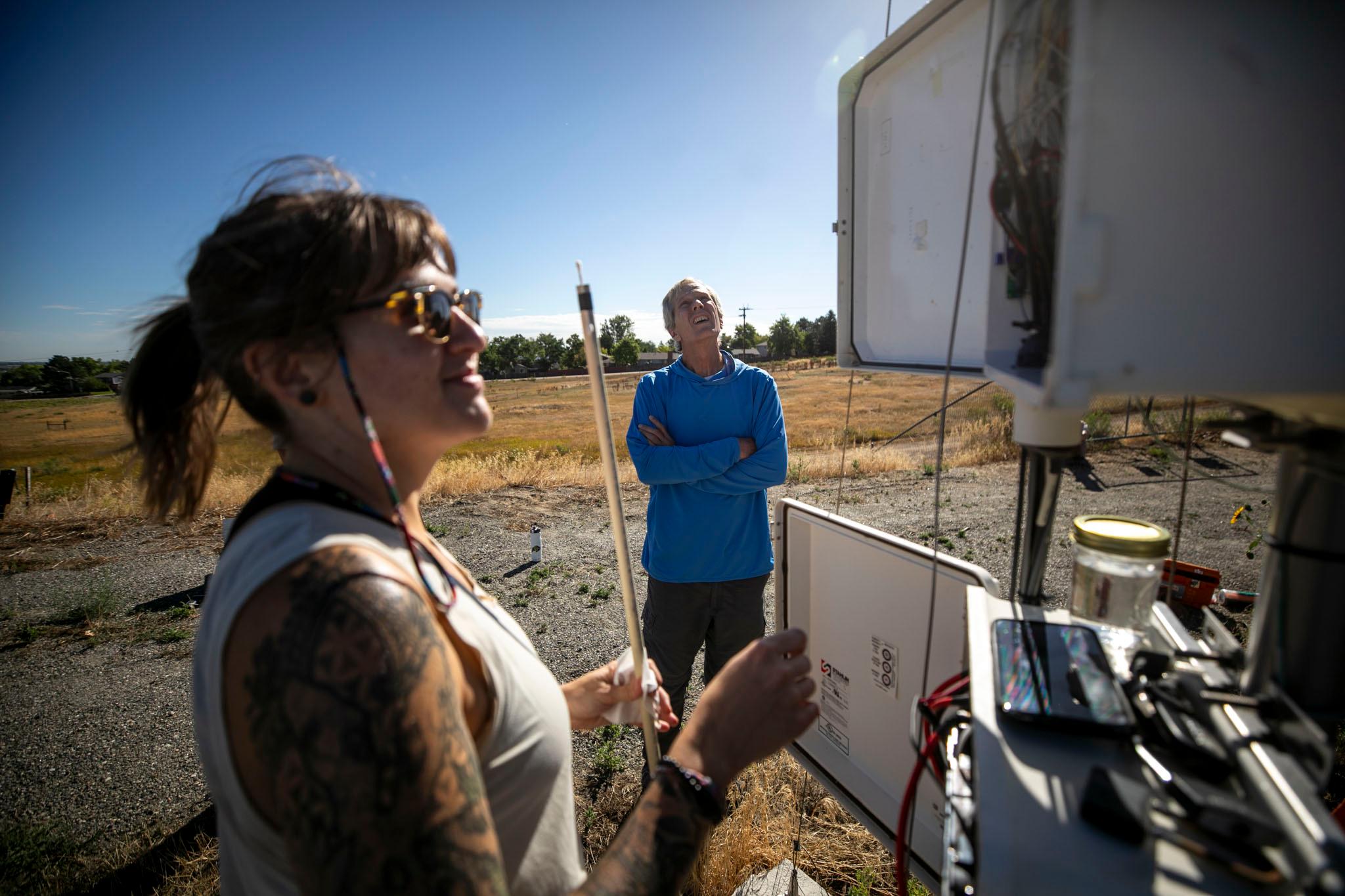 A woman in the foreground looks into an open box, filled with electrical equipment, as a man in the background stares up at the gear.