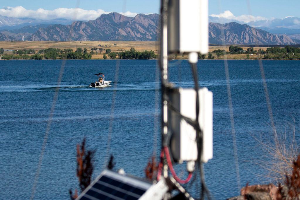 A boat sits on a blue lake, with mountains and green trees rising in the distance; out of focus, in the foreground, is a tower with boxes and solar panels mounted on its side.