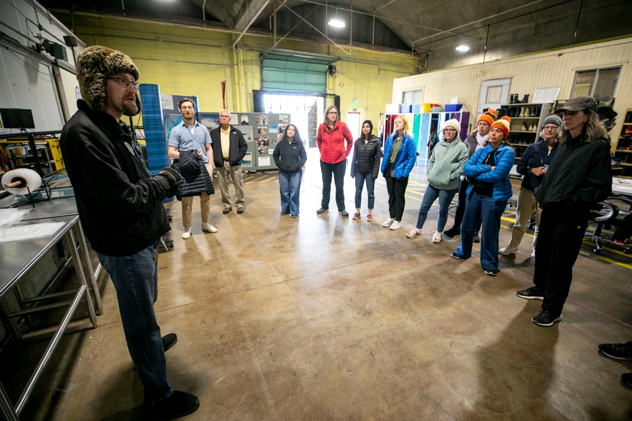 A man in a winter hat speaks to a circle of people bundled up as if for winter, in a warehouse.