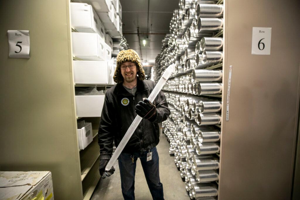 A man in a winter cap, the kind with the fuzzy ears, stands before shelves full of metal tubes. He holds a white cylinder in his hands, which is at least half as tall as he is.