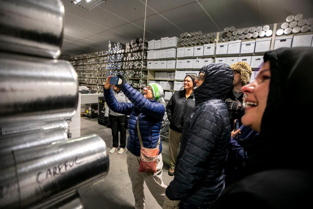 A woman in winter clothing holds up a phone, photographing a shelf full of metal tubes. A woman smiles in the foreground.