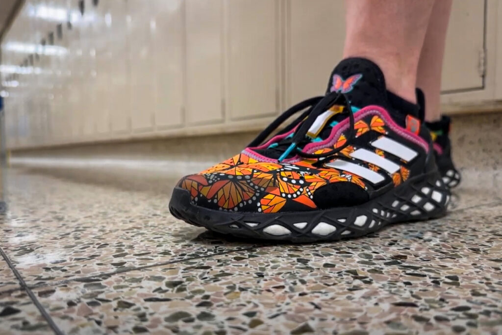 In the foreground is a special Adidas shoe for Dia de Los Muertos, with lockers in the background. The shoe is decorated with orange segments that look like butterfly wings.