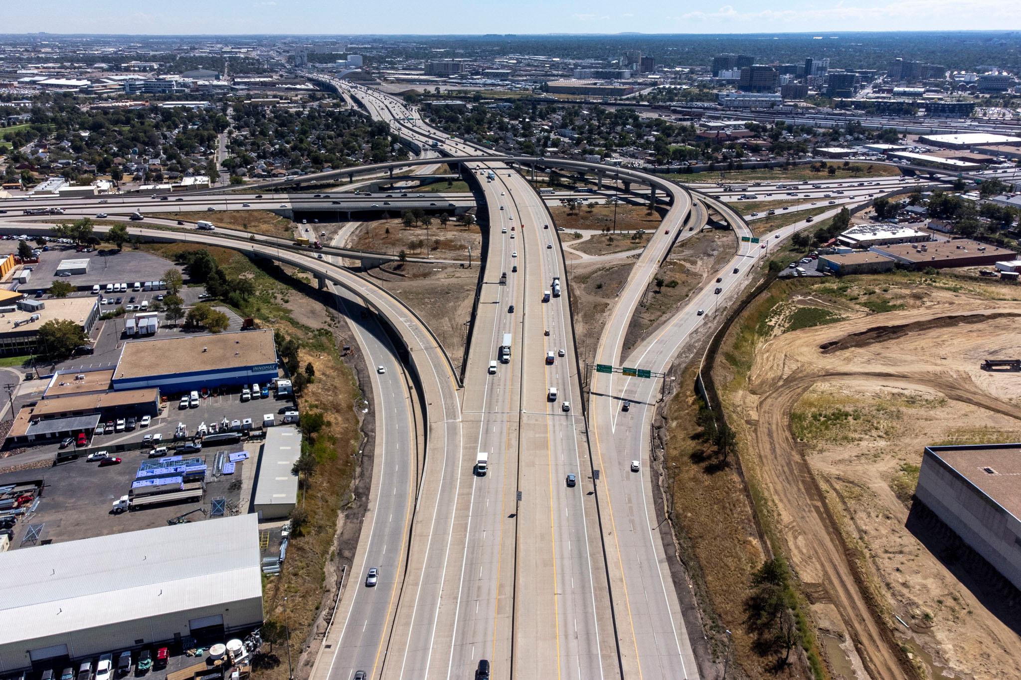 An aerial view of two massive highways joined by a sprawling spaghetti junction. Cars drive along it; the trees nearby are green and the sky is blue.