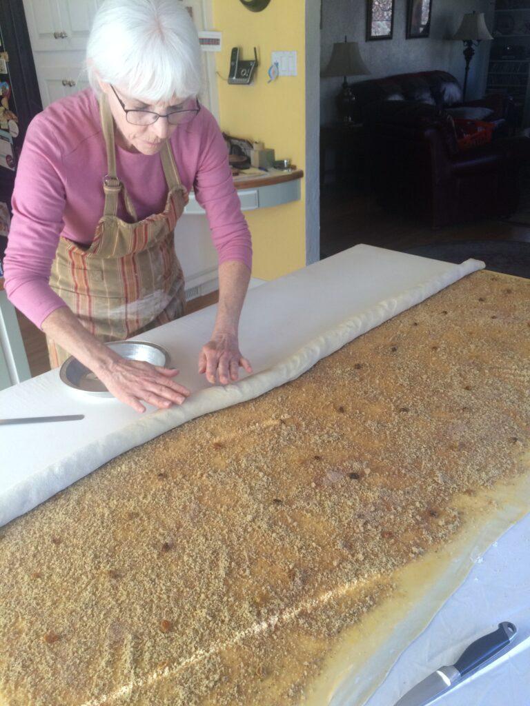 a woman wearing an apron rolls up dough spread with nut filling that covers the entirety of a very long table