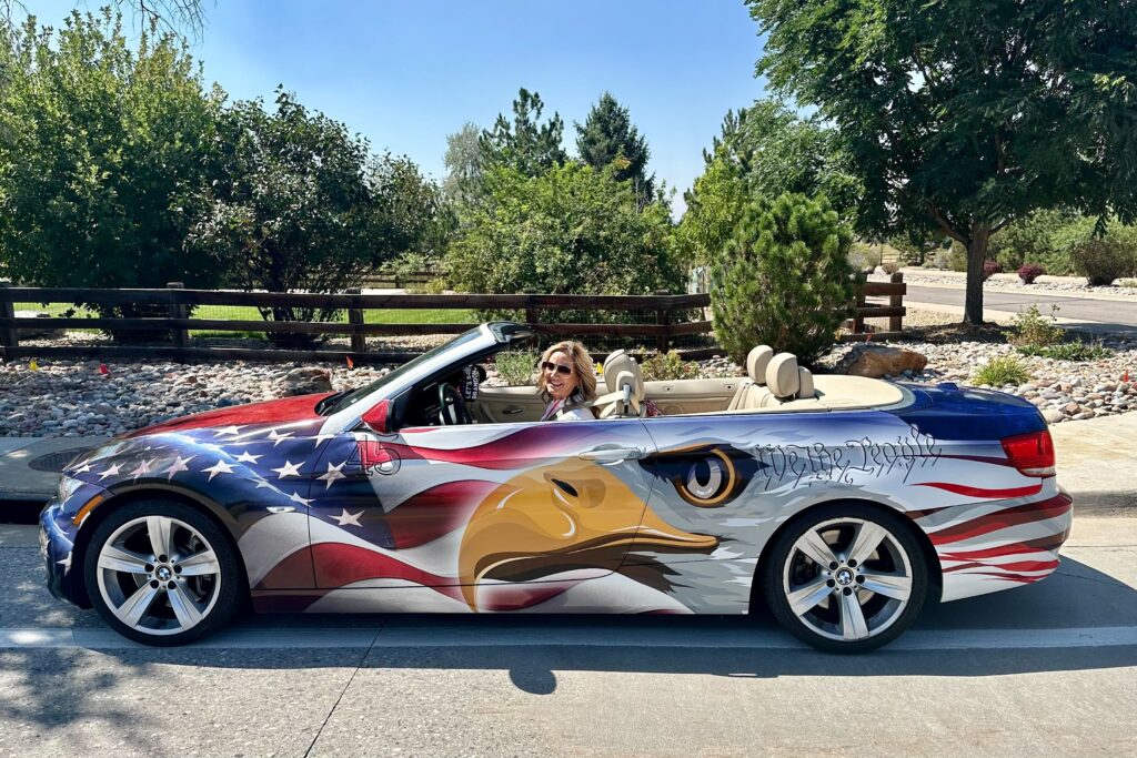 A woman smiles from the driver's seat of a BMW convertible wrapped with an American flag and bald eagle exterior.