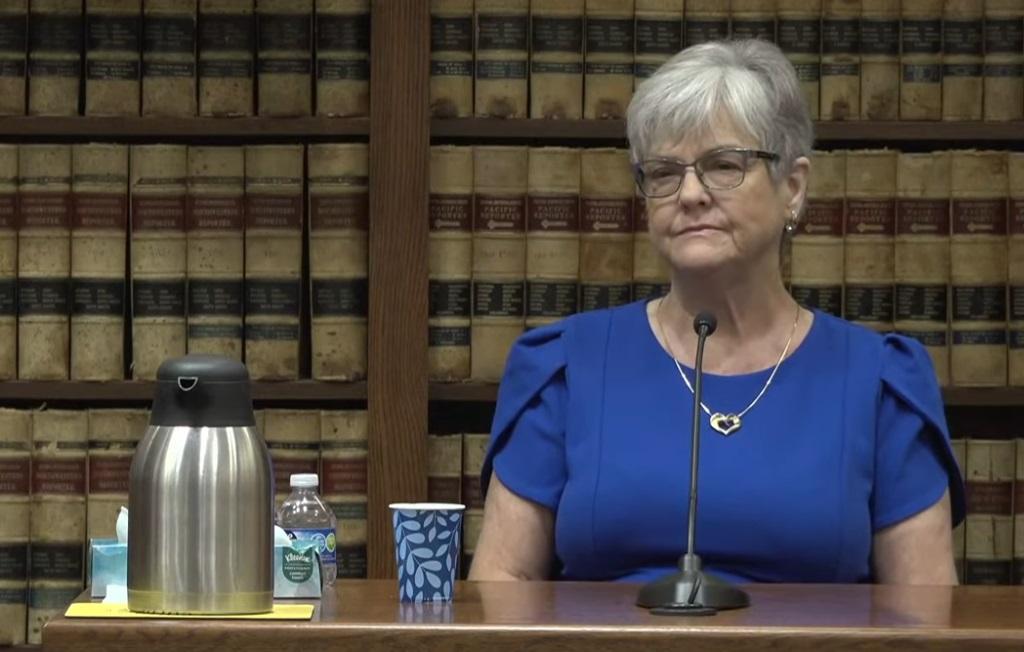 A woman with gray hair, a blue dress and a heart necklace sits at the courtroom witness stand — a brown wooden desk in front of a book shelf filled with law books.