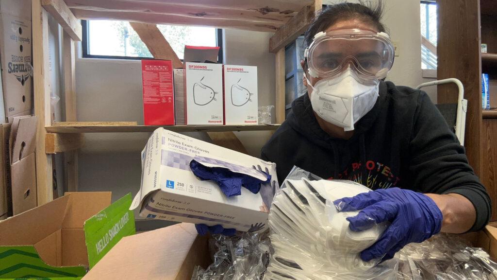 A woman wearing gloves, plastic goggles, and and N95 mask, holds a box of blue nitrile gloves.