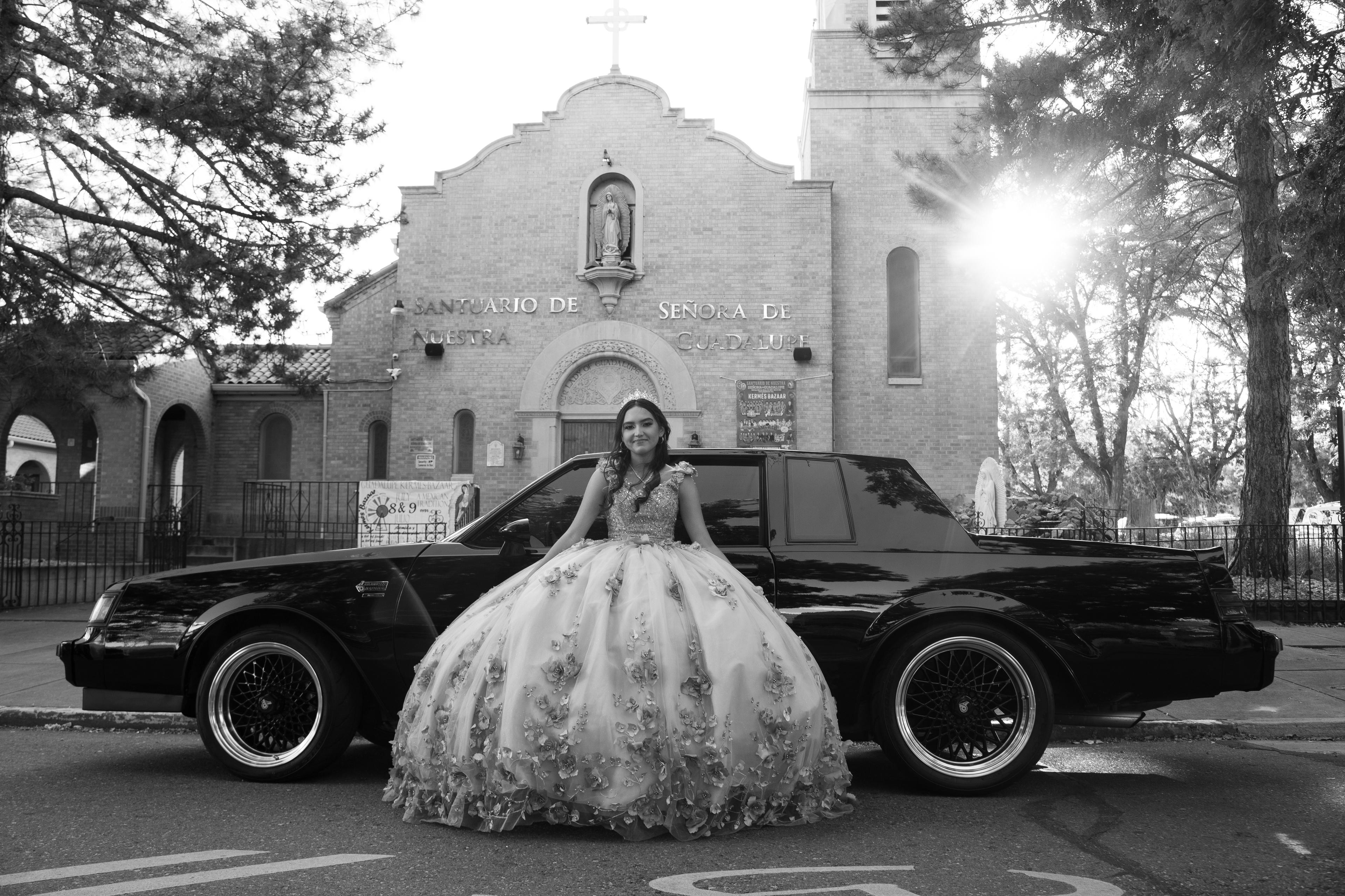 A black-and-white portrait of a girl in a large, floral-embellished quinceañera dress posing in front of a black car, parked on in front of a church.