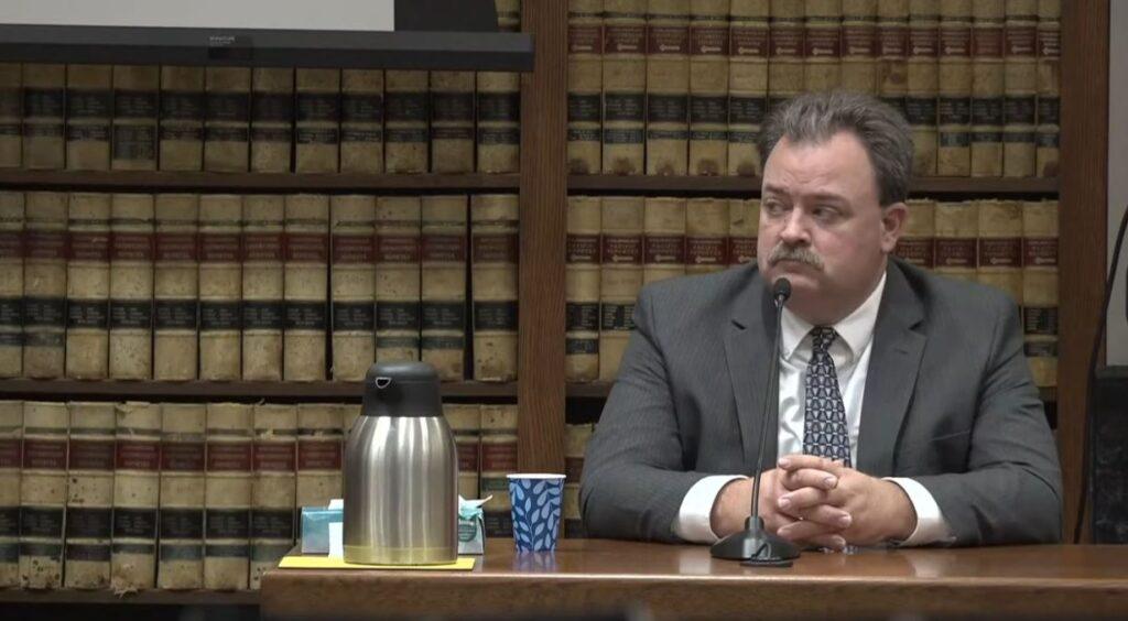 A man named Dallas Schroeder, wearing a gray suit, white shirt and triangle-patterned tie, sits at a wooden table with a microphone in front of him, waiting to testify in a Grand Junction courtroom.