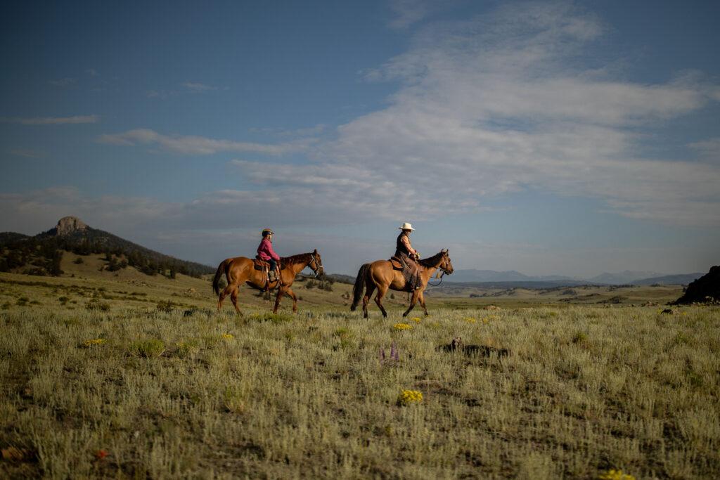 A woman and a child ride across an open grassland with rock formations and mountains in the distance