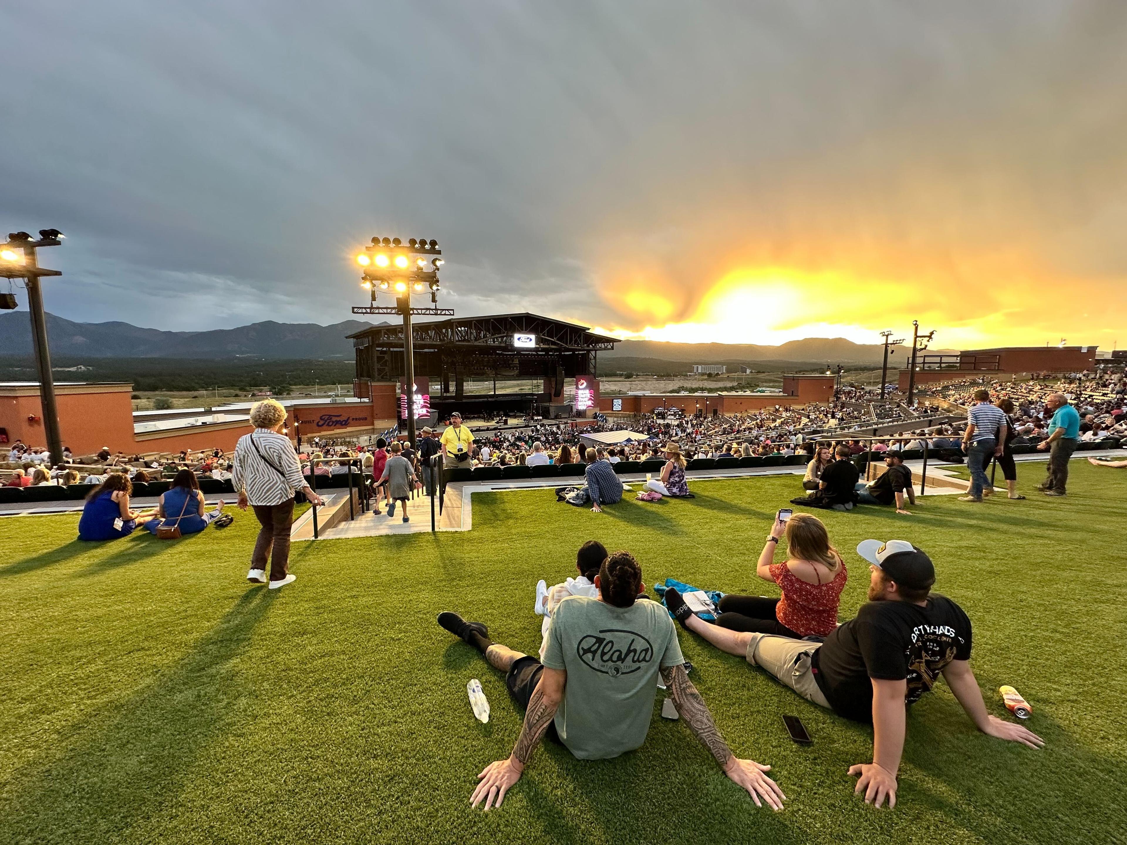 People sit on the lawn at the new Ford Amphitheater in Colorado Springs. They're facing the stage and the mountains. The sun is setting.