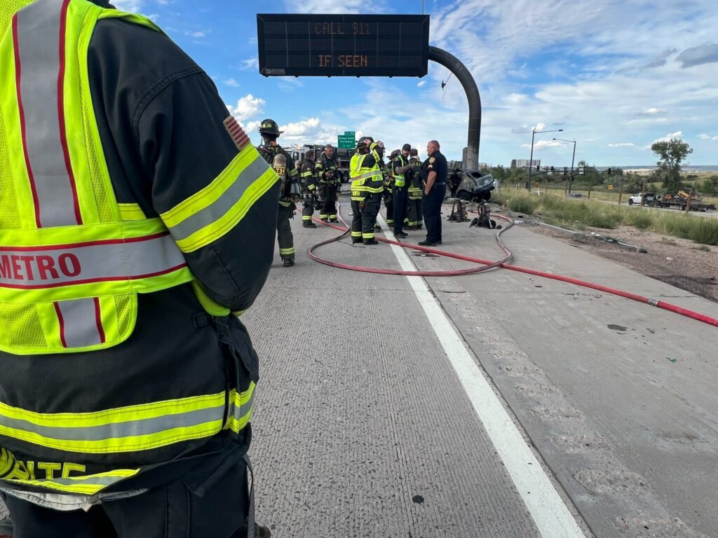 Firefighters on I-25 stand near and around an EV that caught on fire.