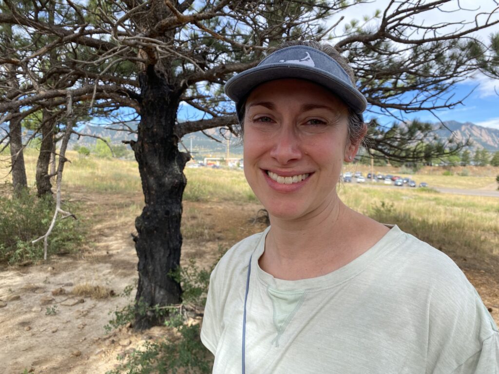 A woman in a blue visor smiles in front of a burned, blackened pine tree.